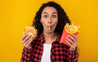 High school woman eating fast food, holding burger and fries, expressive facial expression, happiness, casual clothing, yellow background, fast food cravings, unhealthy eating, junk food.